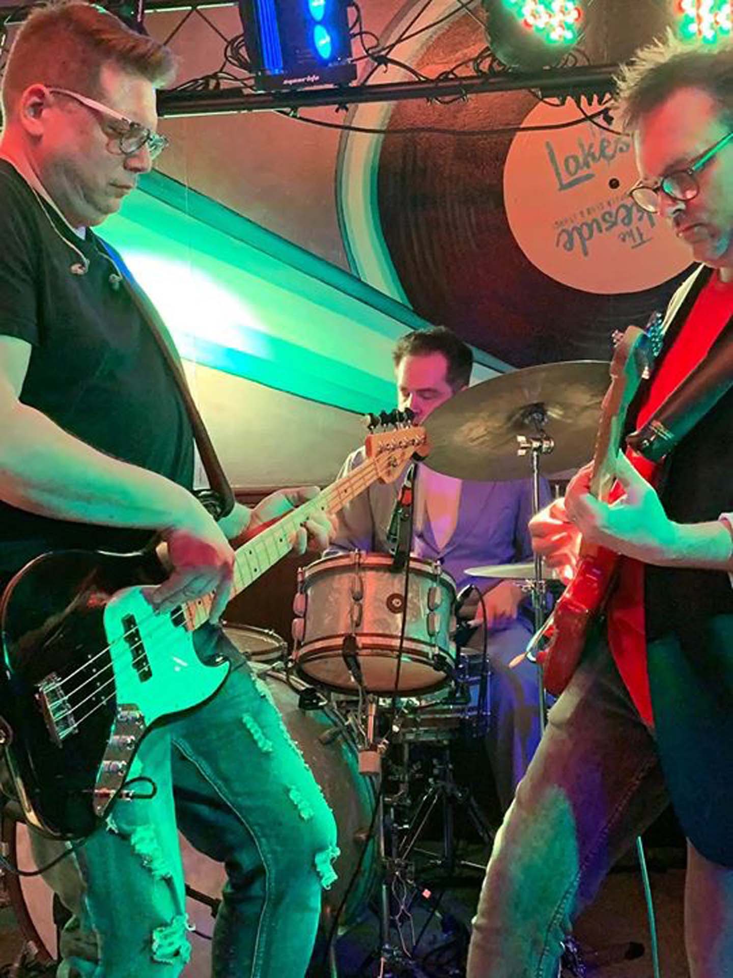 man playing a bass guitar on the left and another playing a guitar on the right with the drummer in the background in the middle.