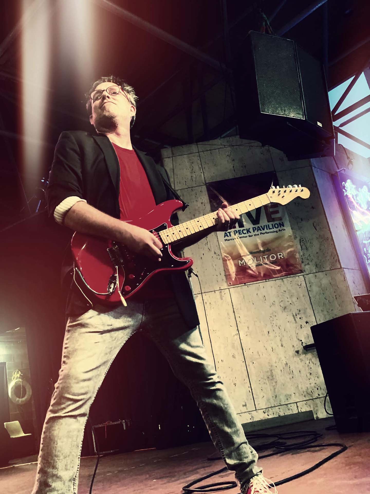 looking up at a guy wearing a red shirt and black suit coat and jeans playing a guitar