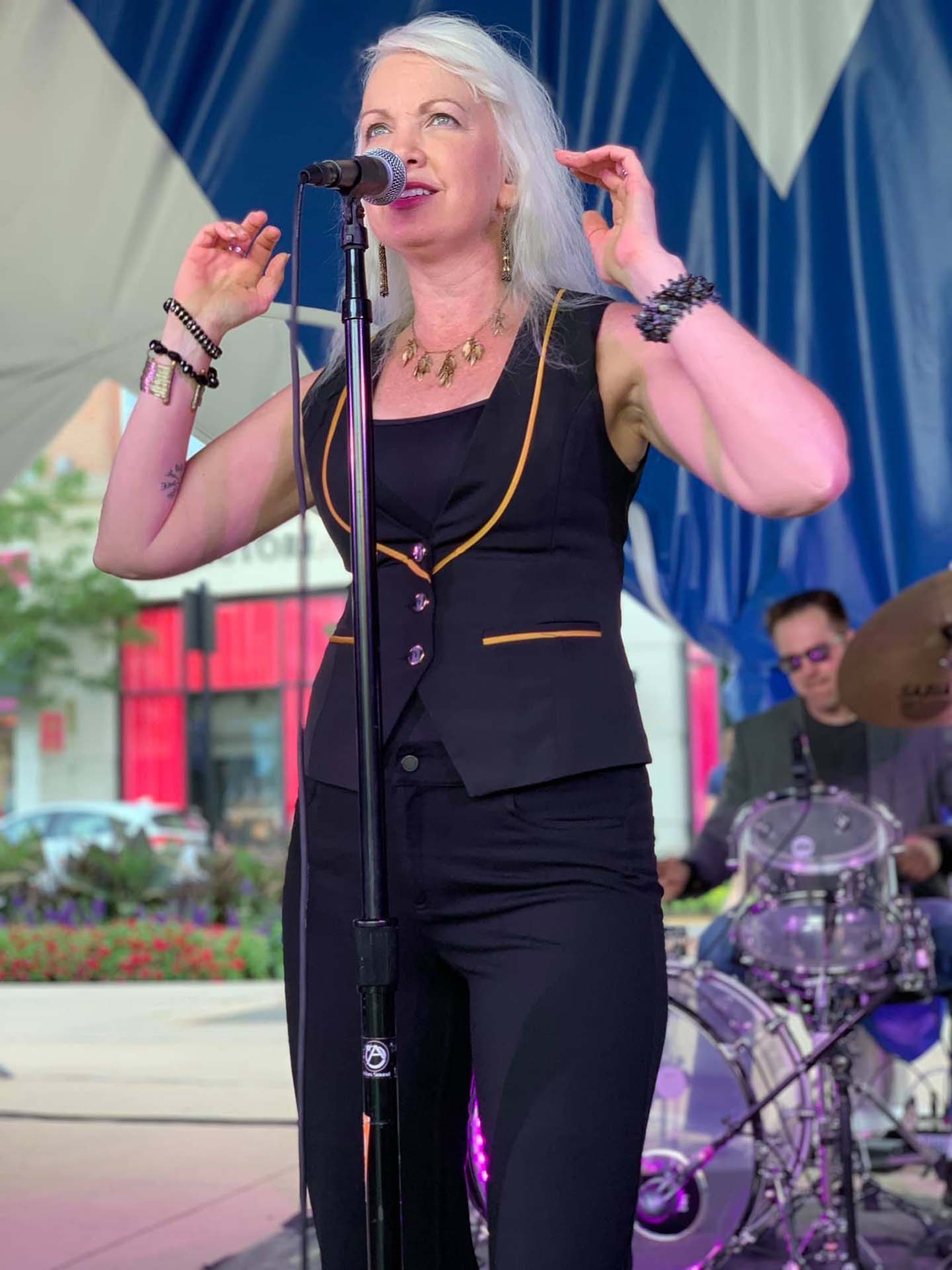 female on stage wearing black suit pants and black and gold trimmed suit vest signing into microphone